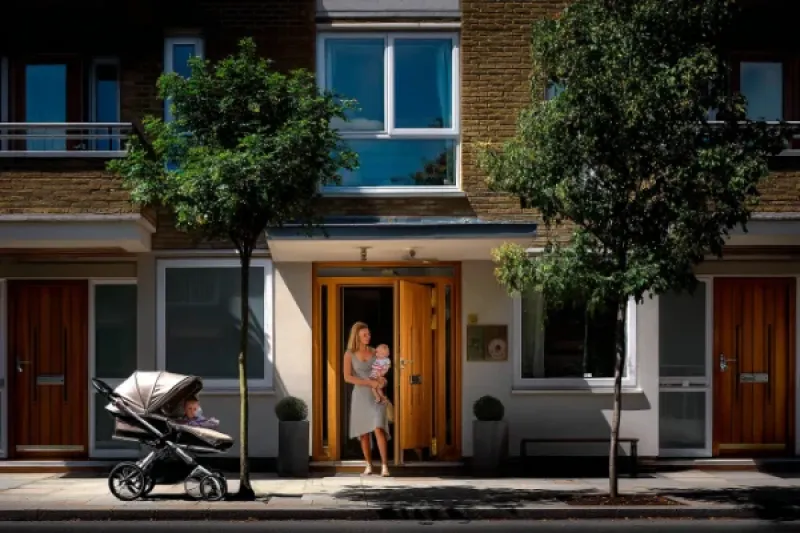 Frau mit Kind auf dem Arm verlässt ein Mehrfamilienhaus. Im Vordergrund steht ein Kinderwagen.